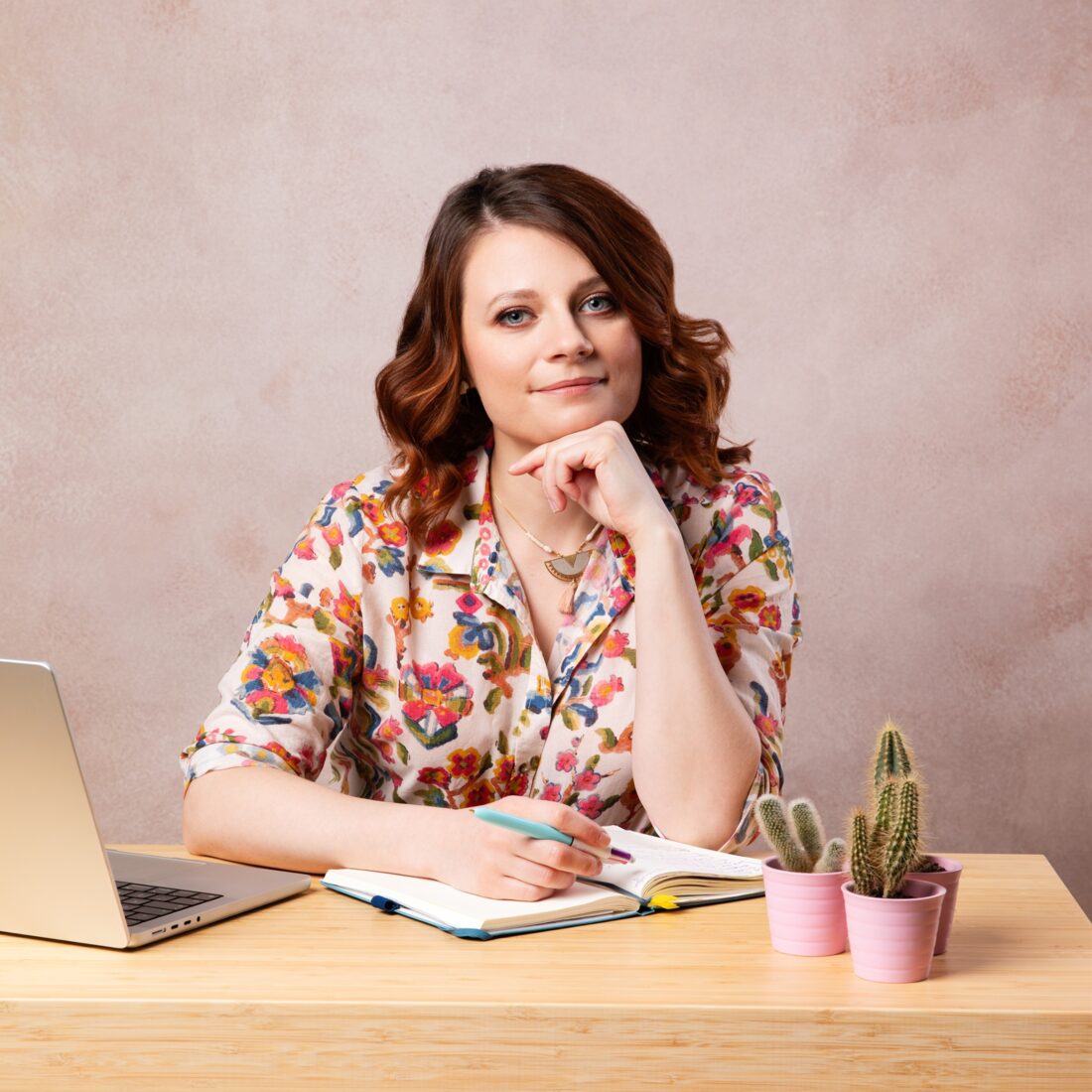Beatka at a wooden desk in a floral shirt, with a laptop, an open notebook and two little potted cacti.